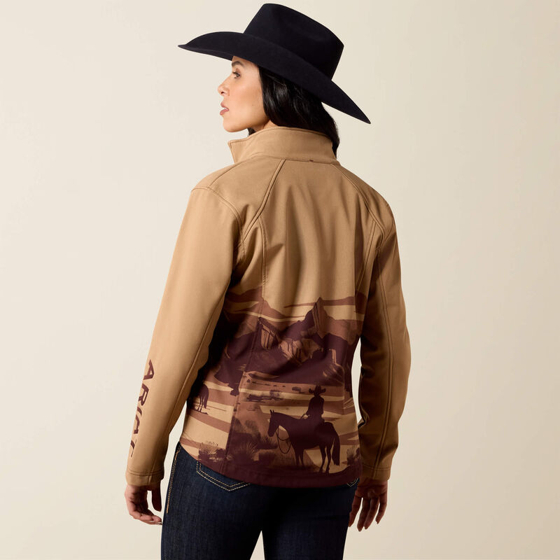 A woman wearing a tan western-style jacket with a desert landscape graphic on the back and a black wide-brim hat, photographed against a plain light beige studio background.