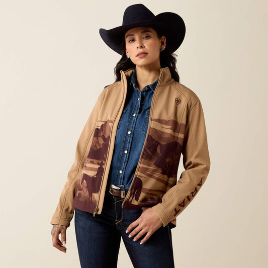 A woman modeling a tan western-style jacket with brown and black landscape print panels on the front and sleeves, worn over a blue denim shirt and jeans, with a black cowboy hat against a plain beige studio background.