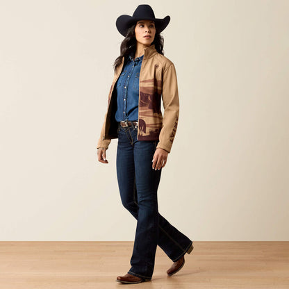 A woman modeling a tan and brown patterned jacket over a blue denim shirt, paired with dark blue jeans, a black cowboy hat, and brown boots, standing against a neutral beige studio background.